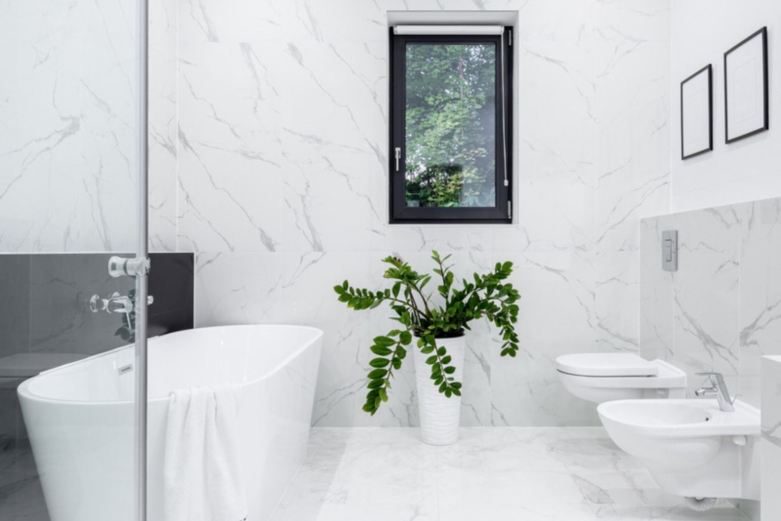 Lyst badeværelse med hvide marmorfliser, fritstående badekar, væghængt toilet, plante og vindue med dagslys